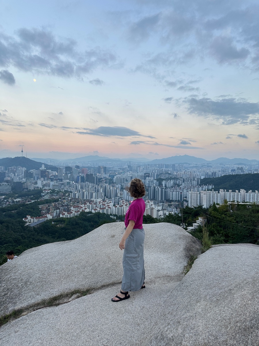 Jenna standing on a granite outcrop above Seoul at dusk, the city skyline and N Seoul Tower behind her